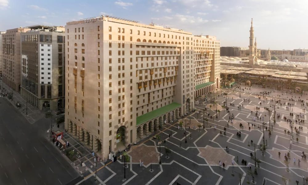 Außenansicht des Sofitel Shahd Al Madinah Hotels bei Tageslicht, mit einem großen Platz und Menschen, die sich bewegen.