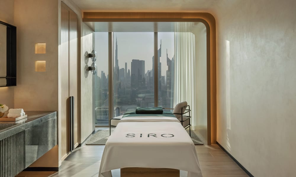 Ein modernes Wellness- oder Behandlungszimmer mit einer Massagebank, großen Fenstern mit Stadt-Skyline und minimalistischer Dekoration.