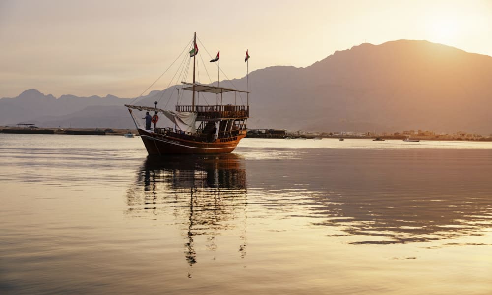 Ein traditionelles Boot schwebt bei Sonnenuntergang auf ruhigem Wasser mit Bergen im Hintergrund.