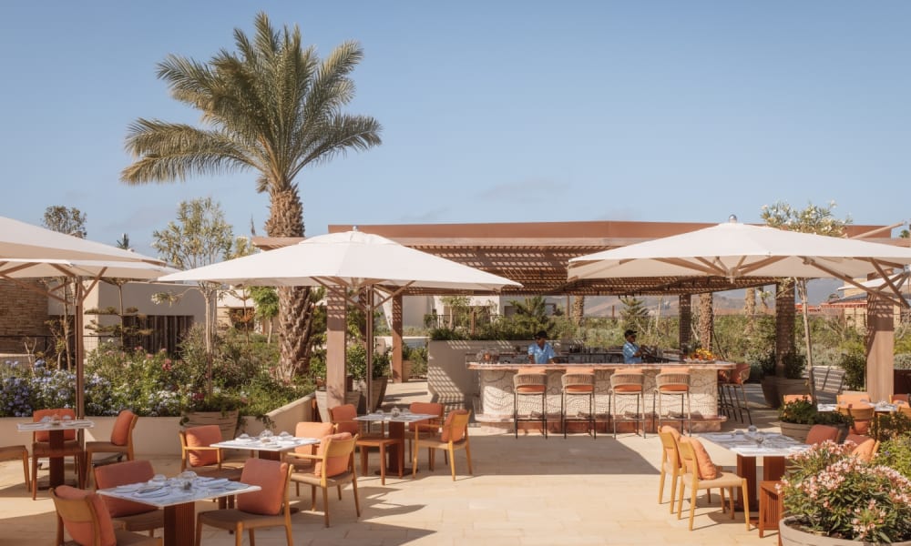 Outdoor-Restaurant mit Tischen, Stühlen, großen Sonnenschirmen, Barbereich und Poolblick bei sonnigem Wetter.