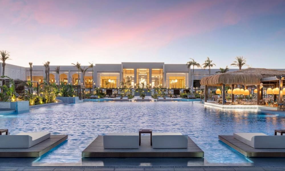 Luxury resort with a large swimming pool, lounge chairs, and a building with glass doors behind palm trees at dusk.