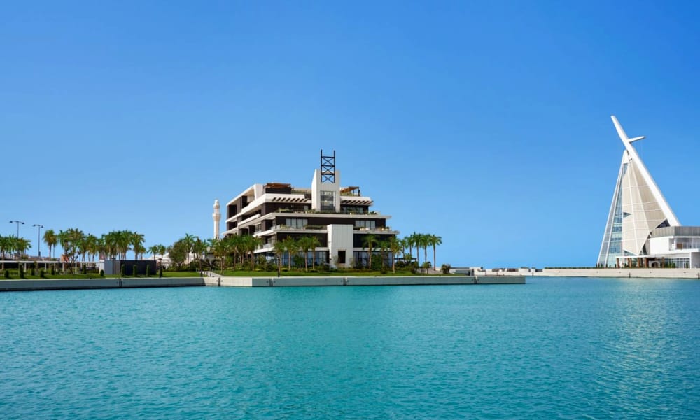 Ein modernes Hotel und ein markantes segelförmiges Gebäude am Wasser, mit klarem blauen Himmel und Wasser im Vordergrund.