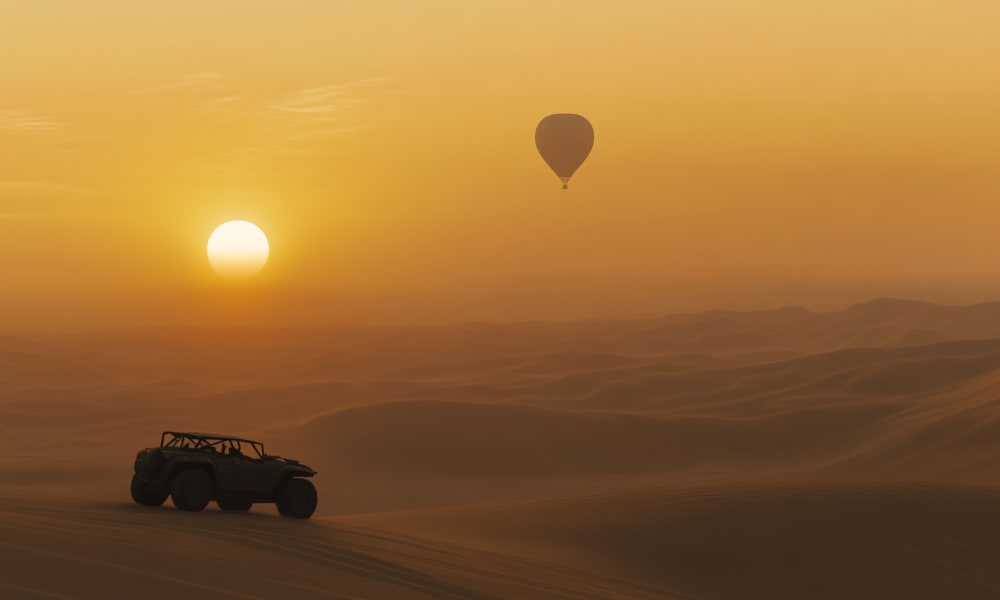 A vehicle driving across sand dunes with a hot air balloon floating in the sky during sunset.