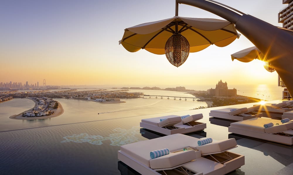 Luxurious outdoor pool area with lounge chairs and umbrellas overlooking a river and cityscape at sunset.