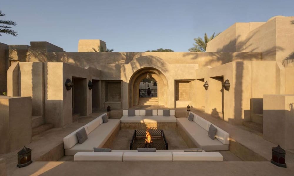 A luxurious outdoor lounge with a central firepit surrounded by comfortable seating, set within a desert-style architecture with sand-colored walls and arches.