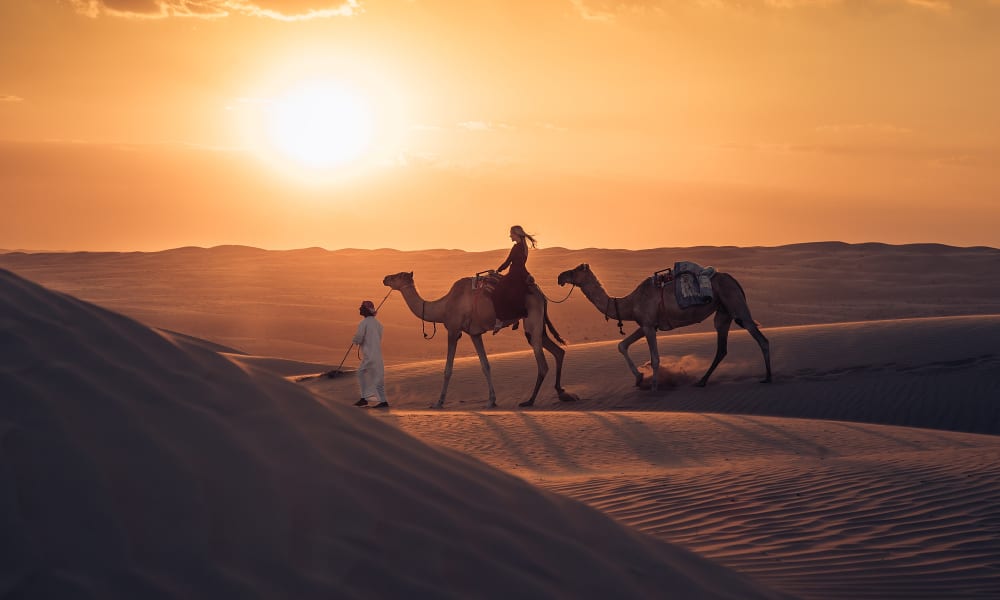 A person leading two camels through sandy desert dunes at sunset, with another person riding one camel, against a vibrant orange sky.