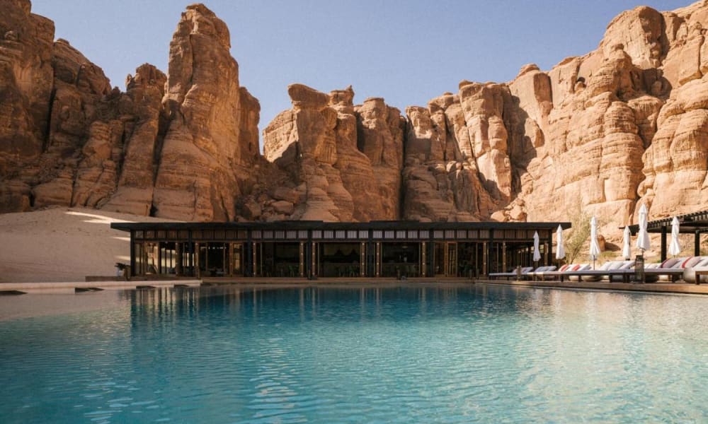 Ein Swimmingpool mit einem modernen Gebäude im Hintergrund, umgeben von felsigem Wüstenland während des Tages.