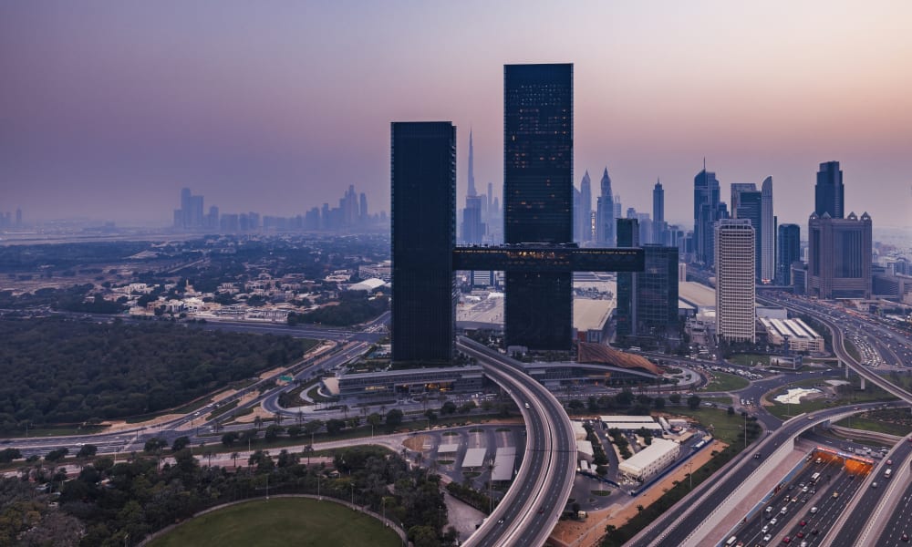 Panoramablick auf eine Stadt bei Dämmerung mit zwei hohen dunklen Wolkenkratzern, die durch eine Himmelbrücke verbunden sind, umgeben von Autobahnen, Grünflächen und zahlreichen anderen Gebäuden.