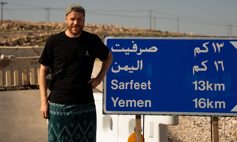 A man stands next to a blue road sign showing directions and distances to Sarfeet and Yemen in both Arabic and English, set in a dry, rocky landscape under a clear sky.
