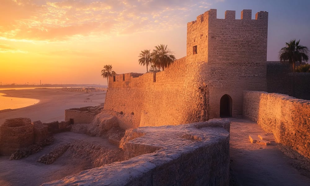 Ancient fortress walls and a tower at sunset, with palm trees nearby, overlooking a beach in Bahrain.