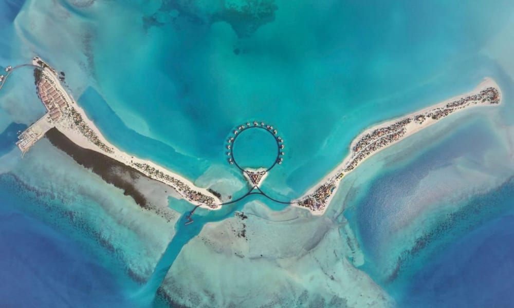 Luftaufnahme eines luxuriösen Resorts mit kreisförmigen Wasserbungalows, die durch eine Brücke verbunden sind, umgeben von türkisfarbenem Wasser und Sandstränden.