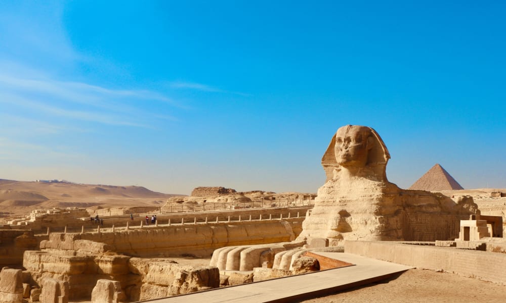 The Great Sphinx of Giza stands prominently against a clear blue sky, with the pyramids visible in the background under bright sunlight.