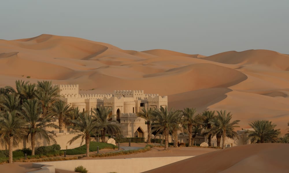 A desert landscape with a historic fortress-style building surrounded by palm trees, set against sand dunes in the background.