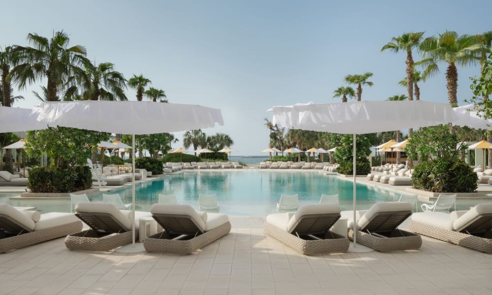Luxuriöse Außenpool-Anlage in einem tropischen Resort mit weißen Liegen und Sonnenschirmen, umgeben von Palmen und klarem Himmel.