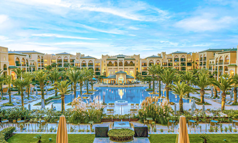 Luxurious resort with a large symmetrical outdoor pool surrounded by palm trees and multi-storey beige buildings under a partly cloudy sky.
