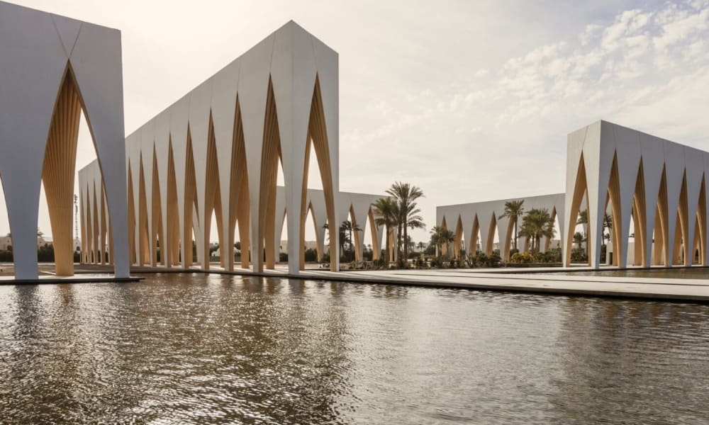 Modern architectural structures with pointed arch designs surrounding a reflective water body, with palm trees visible in the background under a partly cloudy sky.
