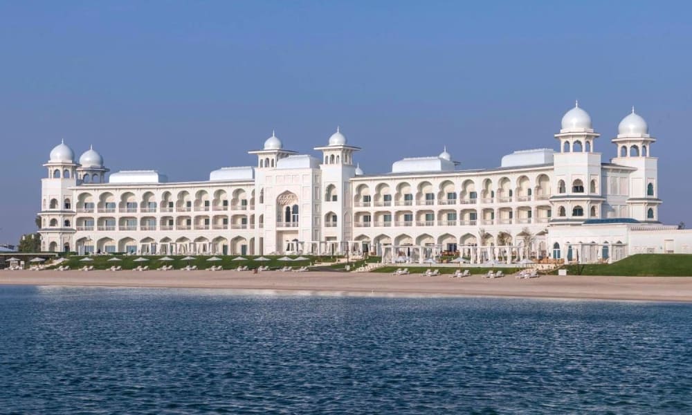 Das Chedi Katara Hotel und Resort, ein großes weißes Gebäude mit Kuppeltürmen, am Wasser gelegen.