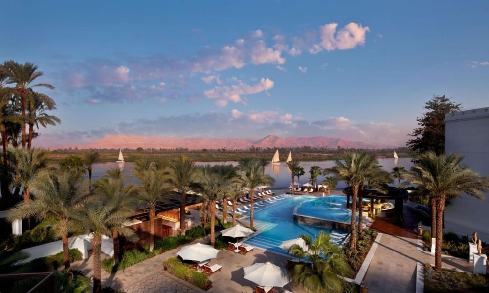 Luftaufnahme des Poolbereichs im Hilton Luxor Resort & Spa mit Palmen und Sonnenliegen, Blick auf einen Fluss und entfernte Berge.