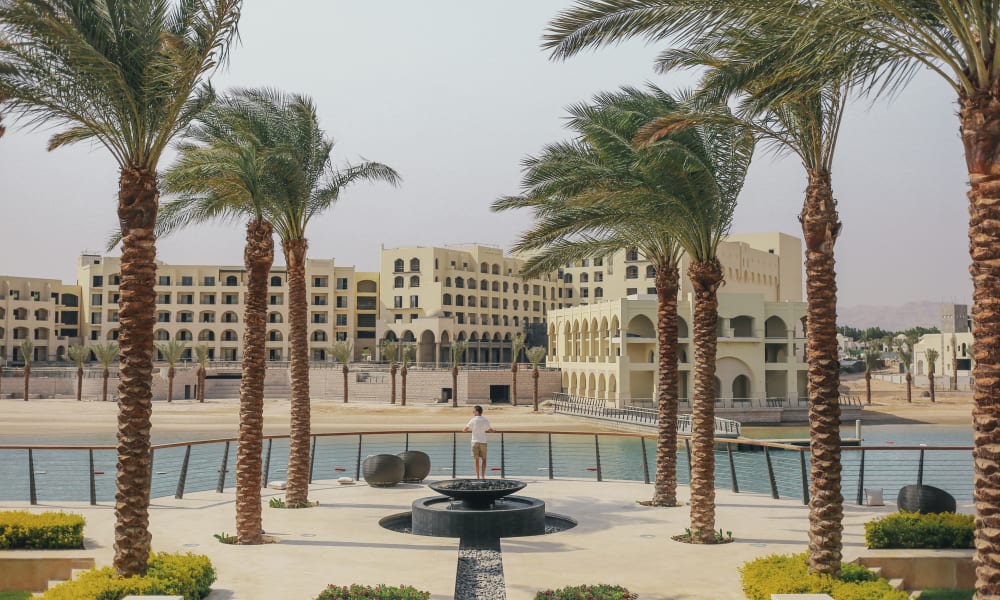 Luxury hotel with multiple arches and palm trees lining a promenade near a pool in a desert setting.