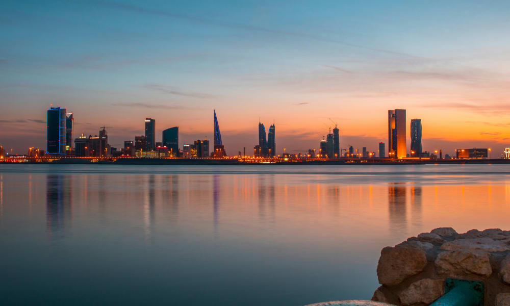 Eine Stadtskyline bei Sonnenuntergang, die auf einem ruhigen Wasser spiegelt, im Vordergrund Steine.