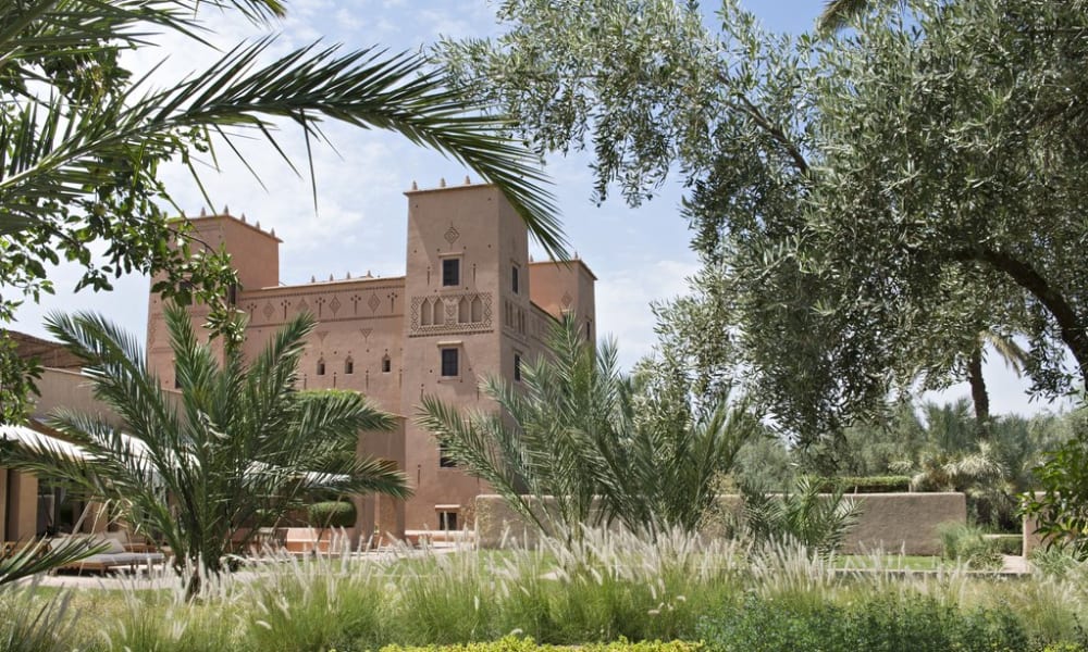 A historic traditional building with a pinkish facade surrounded by lush green trees and plants under a partly cloudy sky.