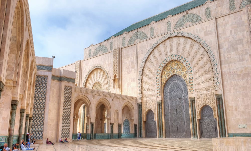 Der große verzierte Eingang einer Moschee in Casablanca mit detaillierten Bögen und dekorativen Fliesen, mit wenigen Menschen, die draußen auf dem Boden sitzen.