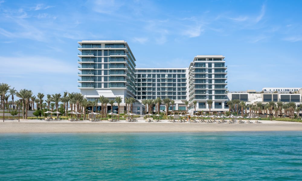 Ein modernes Strandhotel mit mehreren Stockwerken und Glasfassade, gelegen an einem tropischen Strand mit Palmen.