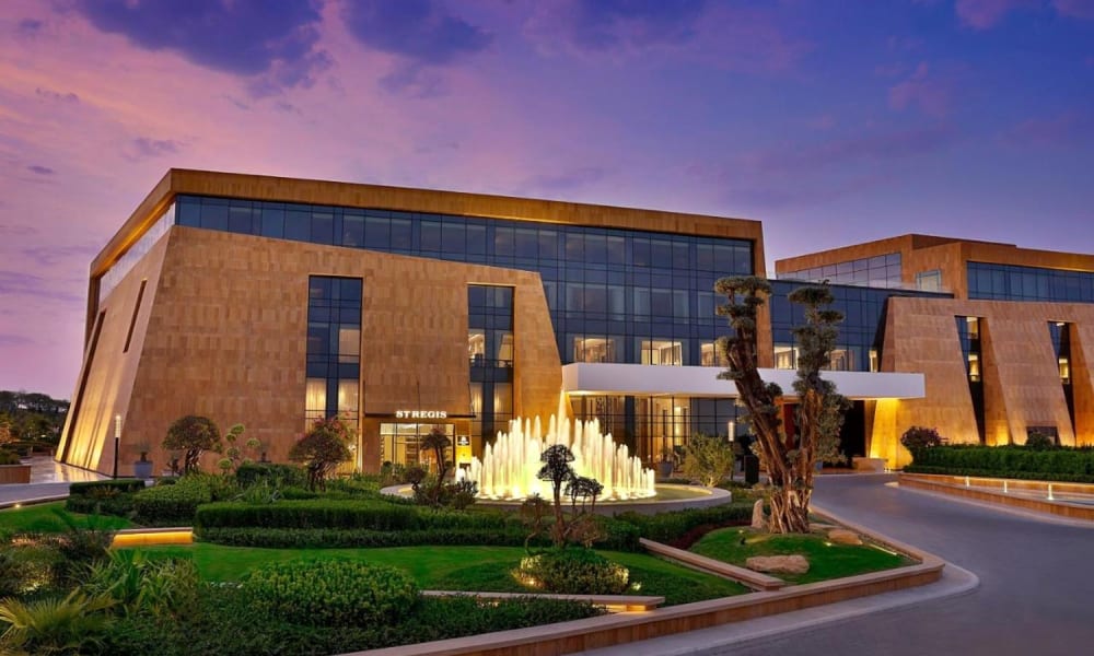 Außenansicht des Hotels St. Regis Riyadh mit beleuchteter Fontäne und gestalteten Gärten bei Dämmerung.
