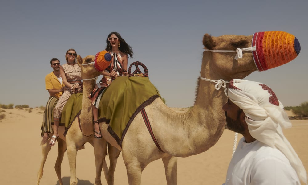 A family riding camels in a desert, with a man in traditional Middle Eastern attire in front of them.