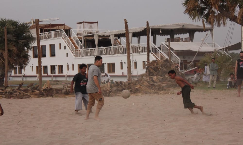 Children playing soccer barefoot on sandy ground near palm trees with a large white building in the background.