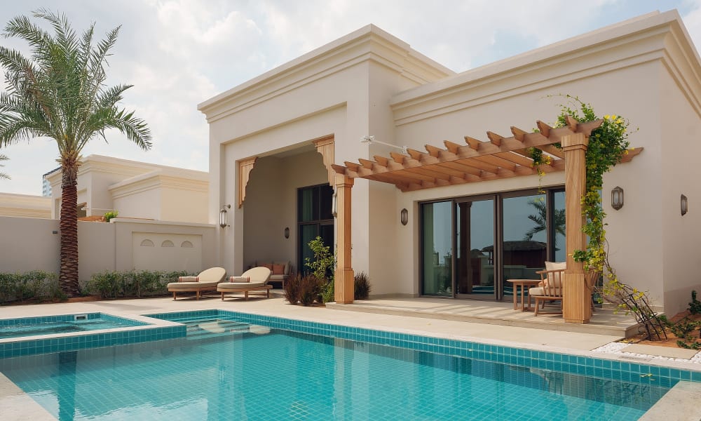 Moderne Villa mit privatem Swimmingpool, Sonnenliegen und Holzpergola, umgeben von Palmen unter bewölktem Himmel.