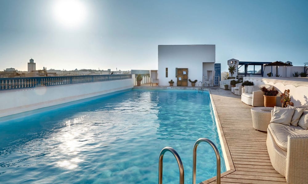 Dachpool auf einer Hotelterrasse mit weißen Sitzbereichen und Stadtblick im Hintergrund unter einem sonnigen Himmel.