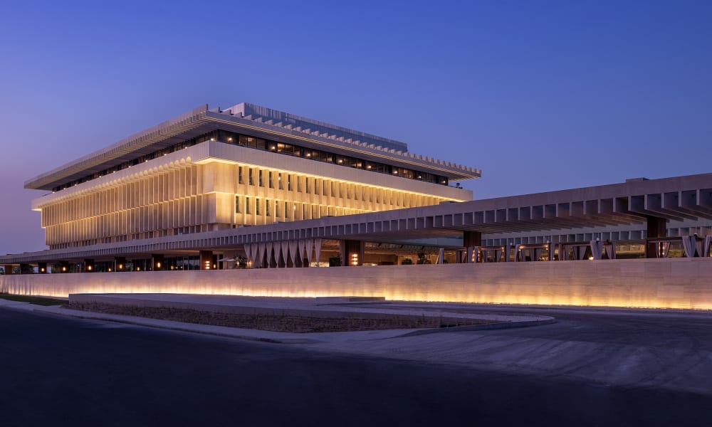 Das The Ned Doha Gebäude bei Dämmerung beleuchtet mit moderner, horizontaler Architektur und warmem gelben Licht.