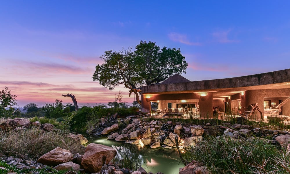 Eine luxuriöse Lodge mit einem Außenbereich, umgeben von Steinen, Wasserstellen und üppigem Grün unter einem farbenfrohen Sonnenuntergangshimmel.