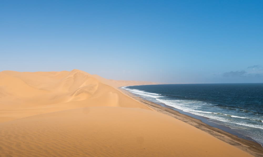 Weite Wüstendünen treffen auf den Ozean bei Sandwich Harbour, Namibia, unter klarem blauen Himmel.