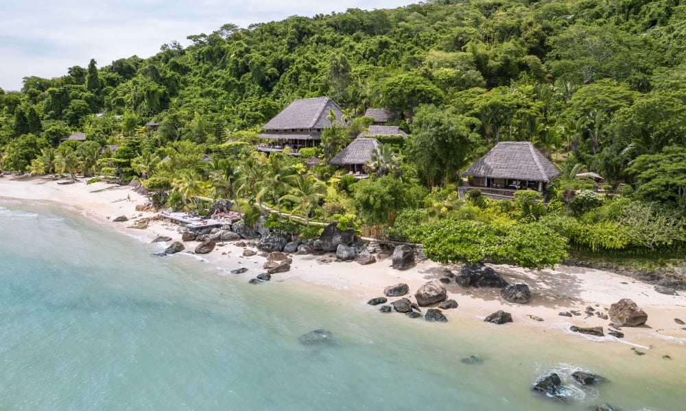 Luftaufnahme von Strandhütten vor üppigem grünen Wald mit Felsen entlang des sandigen Strandes.
