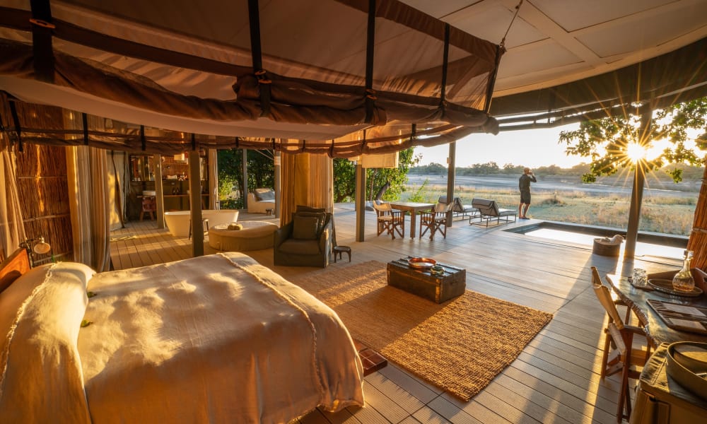 Luxuriöser Safari-Zelt-Innenraum mit großem Bett, Sitzecke und Blick nach draußen auf einen Sonnenuntergang über der Landschaft.