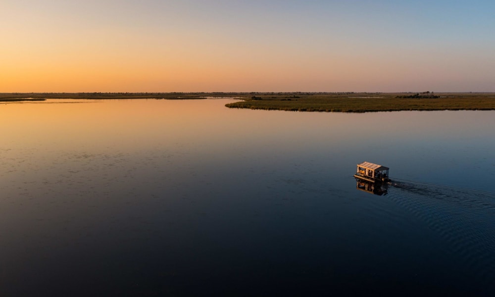 Ein Wasserfahrzeug fährt bei Sonnenuntergang über einen weiten, ruhigen Fluss mit offenen Ebenen im Hintergrund.