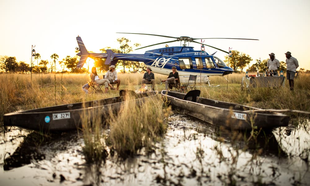 Ein blauer Hubschrauber ist auf einem grasigen Feuchtgebiet gelandet, mit mehreren Personen in der Nähe, zwei Mokoros sind im Wasser im Vordergrund, und die Sonne geht im Hintergrund unter.