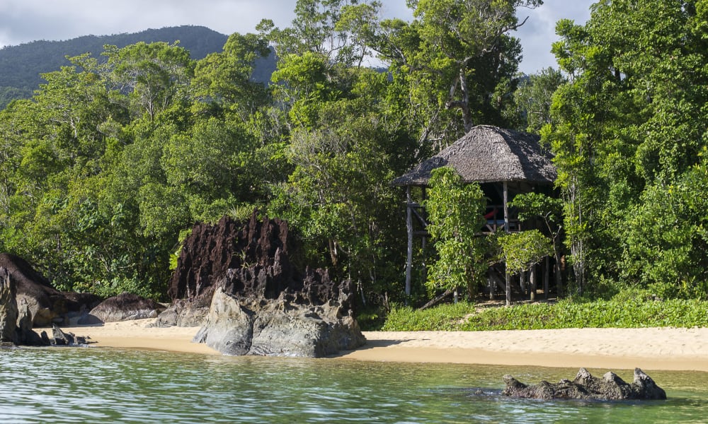 Ein Strand mit einer strohgedeckten Hütte, umgeben von dichtem grünen Wald am Ufer eines ruhigen Gewässers in Madagaskar.