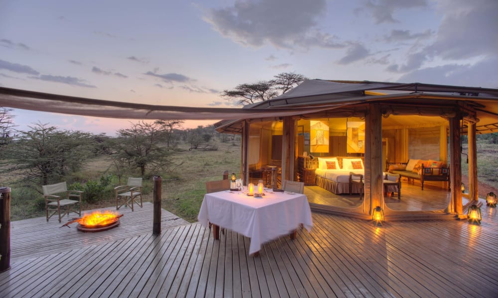 Luxurious outdoor dining area on a wooden deck at sunset, overlooking a savannah landscape, with a tent structure illuminated from within.