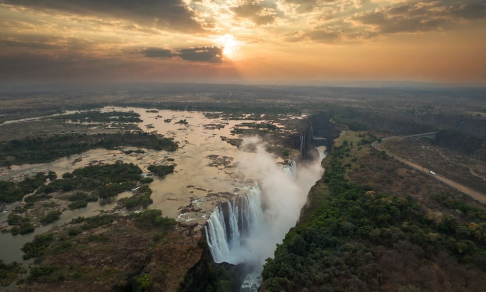 Luftaufnahme der Victoriafälle bei Sonnenuntergang mit der Sonne, die teilweise hinter Wolken verborgen ist und den Wasserfall sowie die umliegende Landschaft beleuchtet.