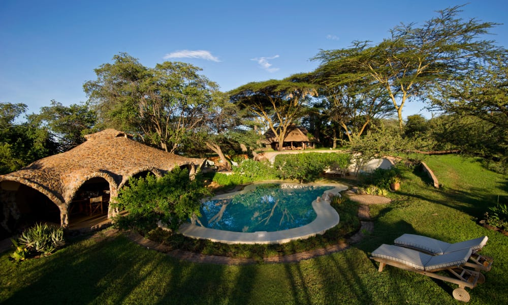 A scenic view of a garden with a round swimming pool, lush green trees, and thatched-roof structures under a clear blue sky.