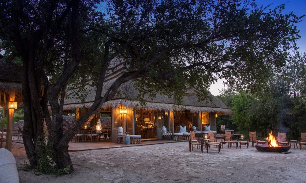 A cozy outdoor lounge area with a thatched-roof building, illuminated by warm lights, and a campfire burning on the sandy ground, surrounded by trees.