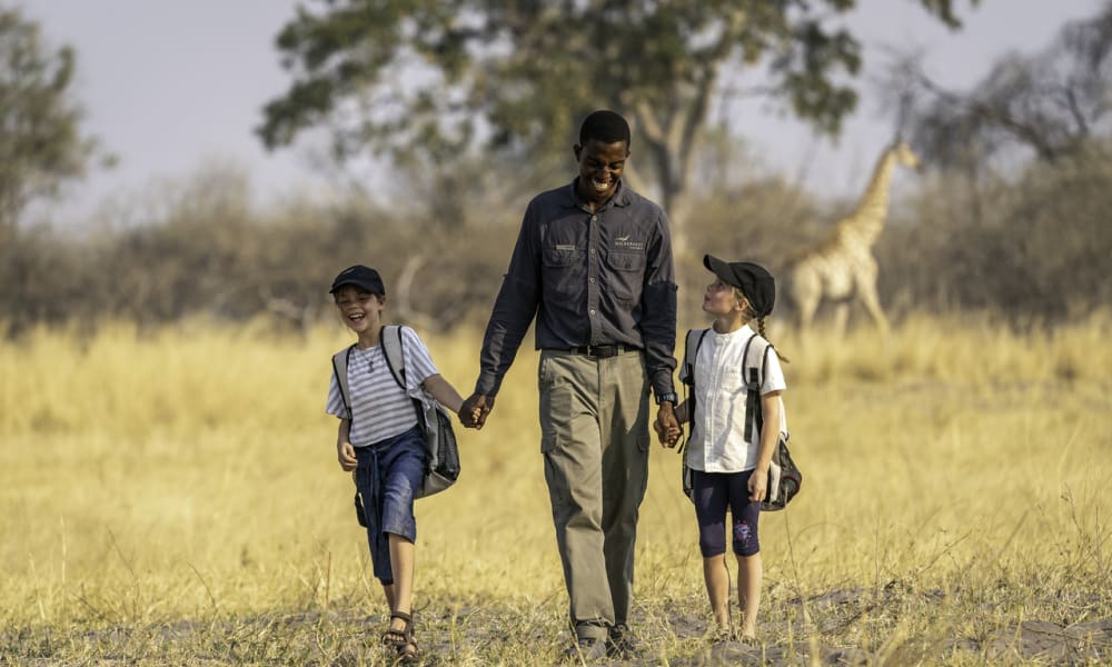 A man walking hand in hand with two children in a grassy savannah with a giraffe in the background.