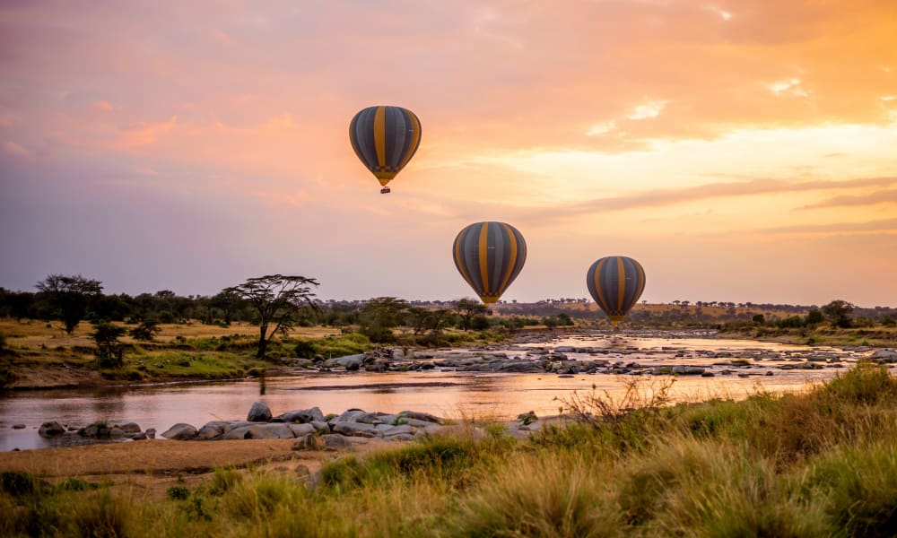 Drei Heißluftballons schweben bei Sonnenuntergang über den Serengeti-Ebenen mit Bäumen und einem Fluss darunter.
