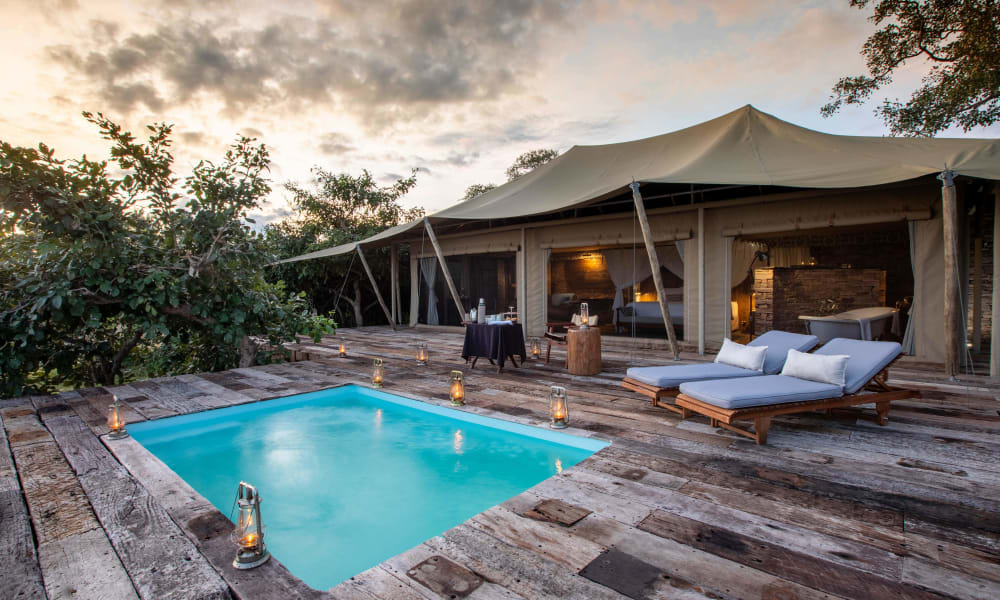 Eine luxuriöse Außensuite mit kleinem Swimmingpool auf einer Holzterrasse, umgeben von Natur, bei bewölktem Abendhimmel.