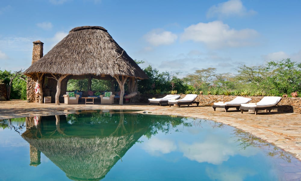 A tranquil swimming pool with a thatched-roof hut and lounge chairs beside it, set in a sunny outdoor location.