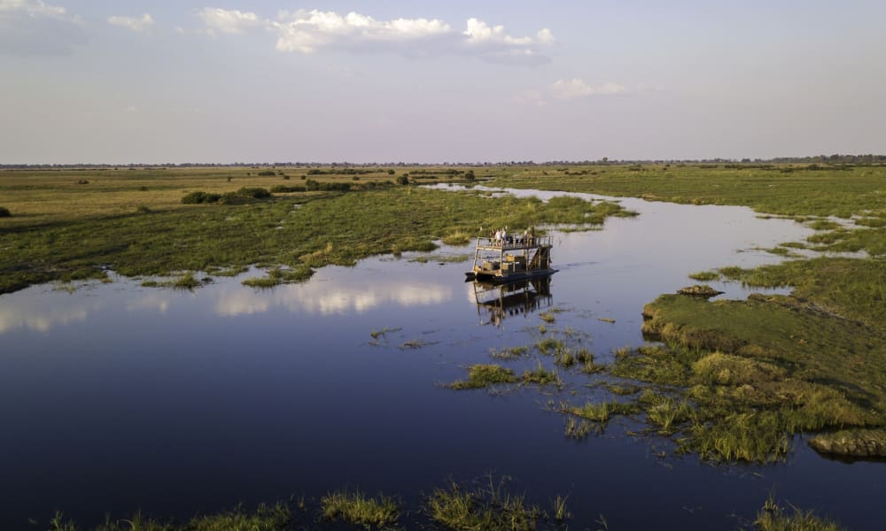 A boat navigates through a wide, calm river amid lush green wetlands under a partly cloudy sky.