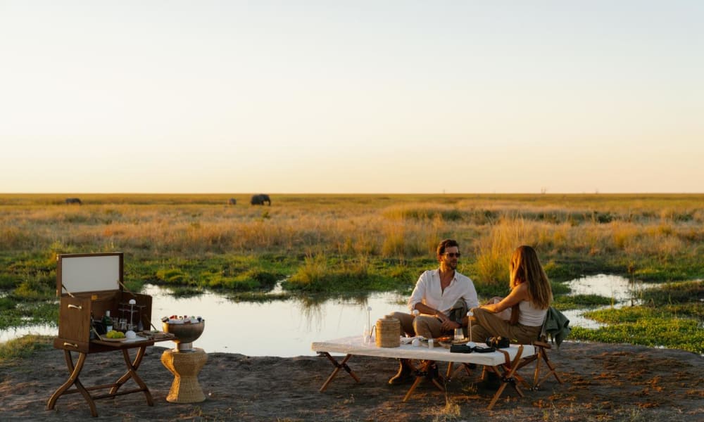A couple having a picnic on the riverbank during sunset in a flat grassy landscape.